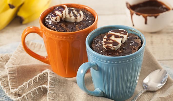 Fotografía en tonos beige, azul y naranja de una encimera blanca con un paño beige, sobre ella una taza azul y una taza naranja con pastel de taza. En el fondo un racimo de plátanos y un recipiente redondo con almíbar.