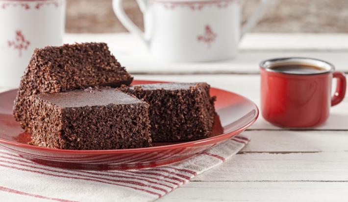 Fotografía en tonos rojos sobre una encimera de madera clara con un paño rayado en blanco y rojo y un plato rojo con tres piezas de bizcocho de cacao con coco. A su lado, una taza de café roja y al fondo, dos teteras blancas.
